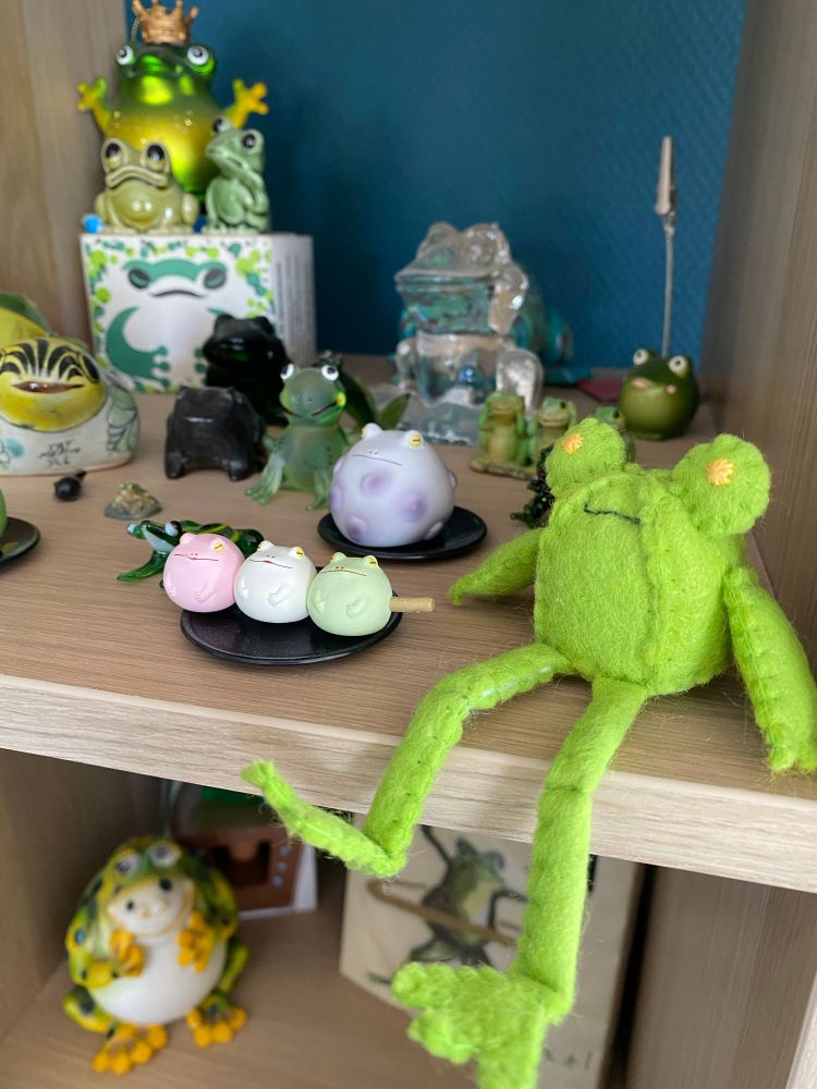 Photo of floris the frog without his yellow raincoat felt doll on a shelf amongst other frog figures and glass trinkets