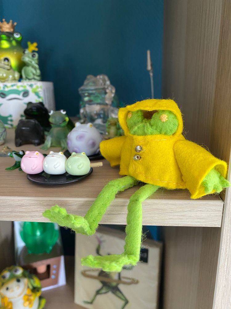 Photo of floris the frog in his yellow raincoat (hood up) felt doll on a shelf amongst other frog figures and glass trinkets
