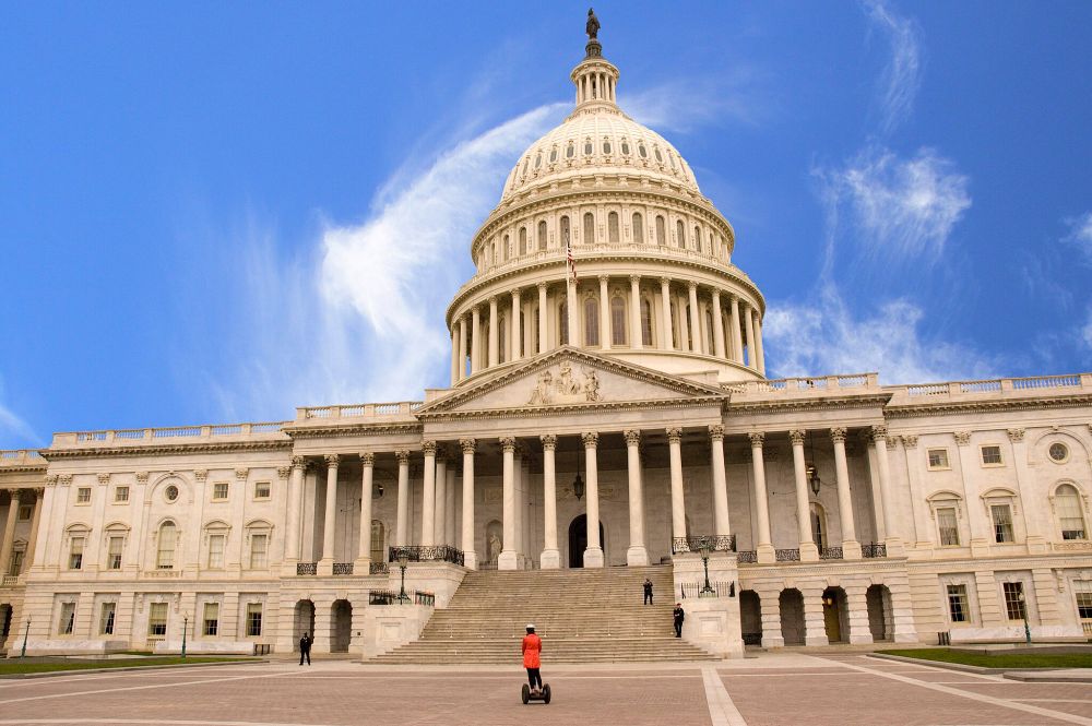 Person in a red coat on a Segway in front of the U.S. Capitol building against a blue sky with whispy white clouds