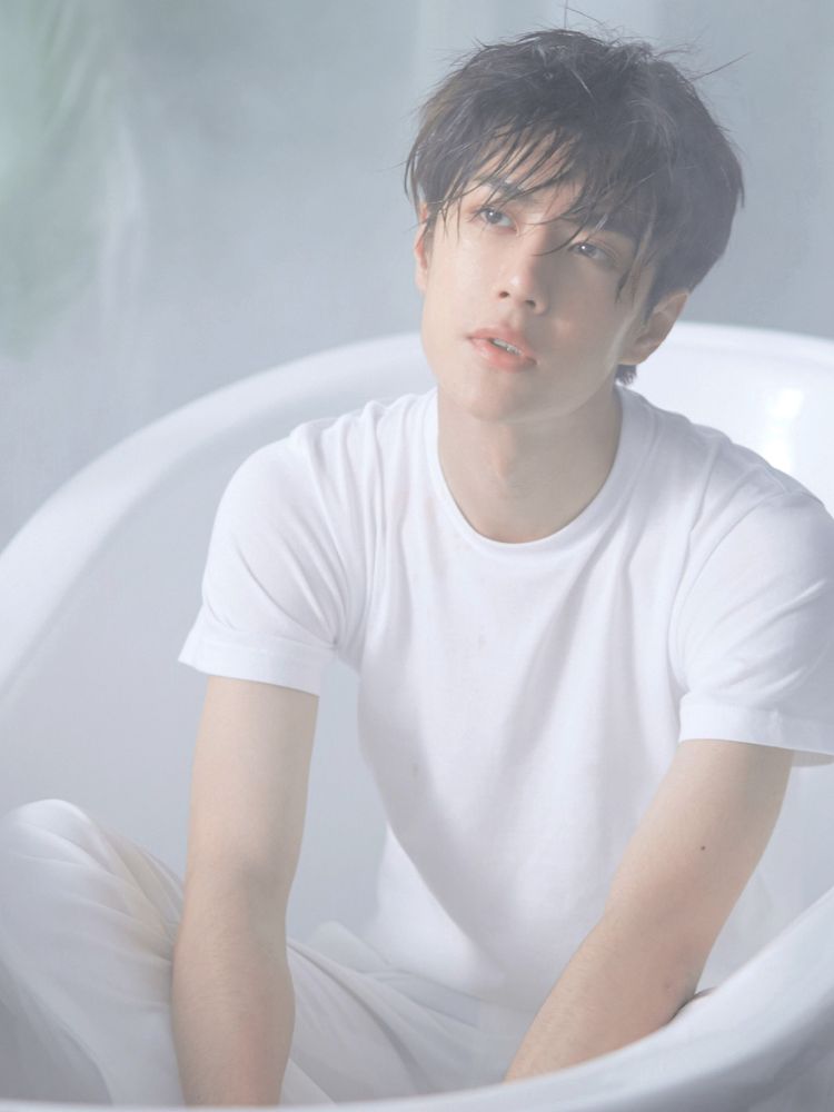 Chinese dancer, singer, and actor Wang Yibo, dressed in a white t-shirt and white jeans, sits in and empty white bathtub. His hair is damp, his skin dewy, and he looks up and to the right, while slightly biting his lip.