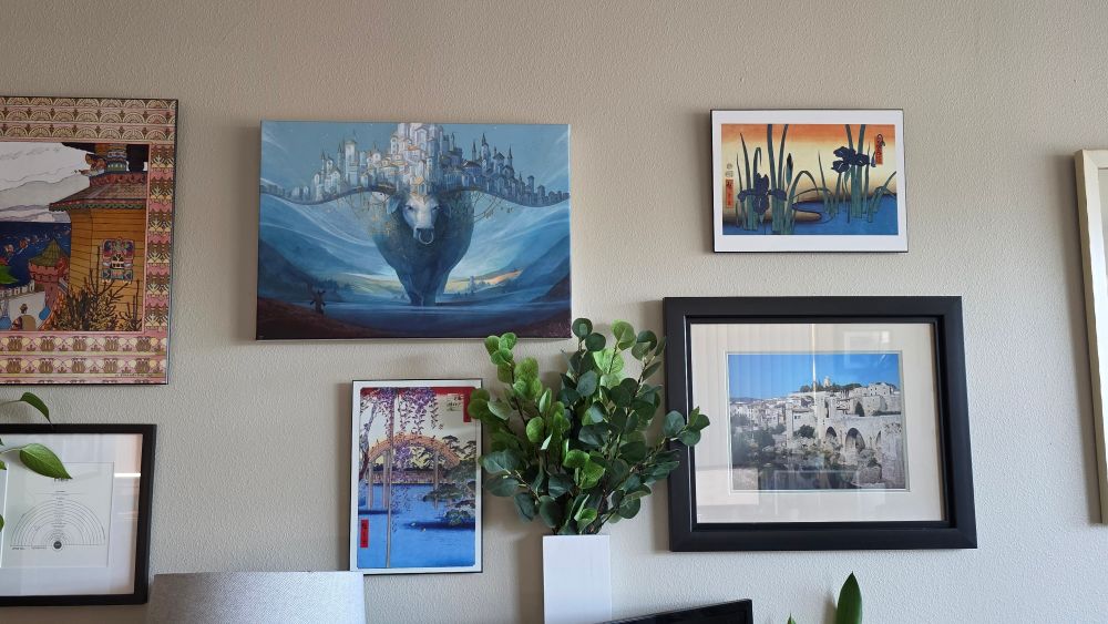 A living room wall decorated with art prints and large framed photos. At the center is OP's glorious ox print featuring a massive ox with a city built across its horns, walking through a valley at sunset in moody, atmospheric colors, predominantly blue.