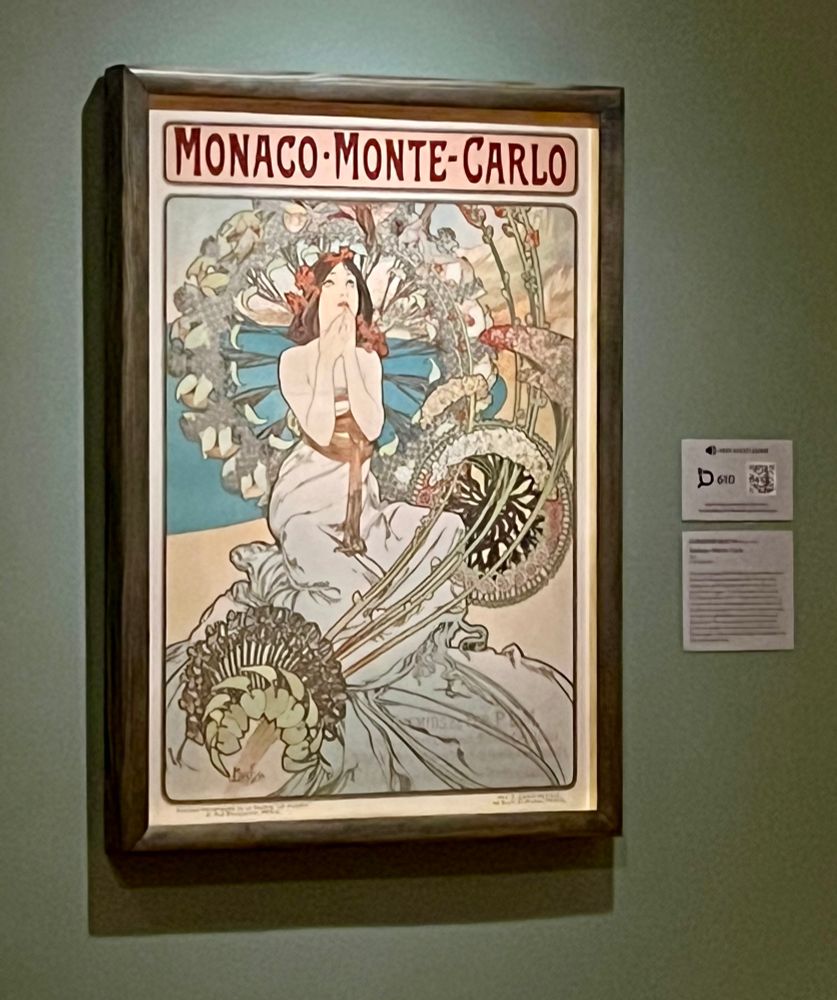 A poster by Alphonse Mucha advertising Monaco and Monte Carlo, with a woman in a white dress and red hair in the center, wrapped in fantastic swirls of vines. 
