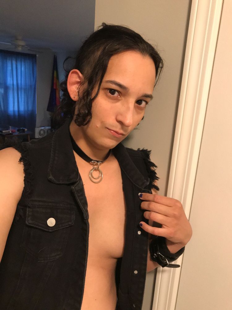Selfie of Rosie Sanchez. She wears a black denim vest with nothing underneath, a thick black wrist cuff, and thin black leather collar with a dual ring lead. She is holding open her vest just enough to see the beginning of her breast and she is looking suggestively into the camera.