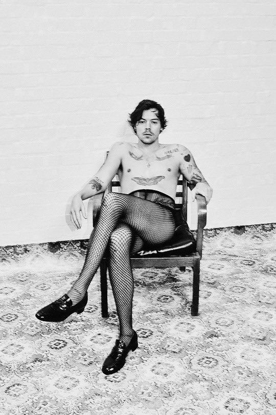 Black and white photo of Harry Styles wearing fishnet tights, black loafers, and sitting in a chair with his legs crossed. 