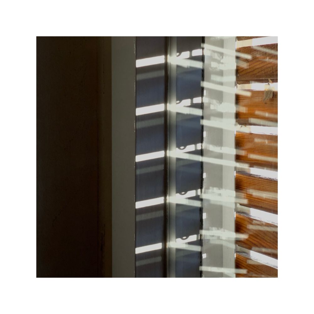an angled closeup view at a window frame. the wooden shutters are closed but small gaps let some stripes of light still through. the incoming light is partly reflected on the two layers of glass