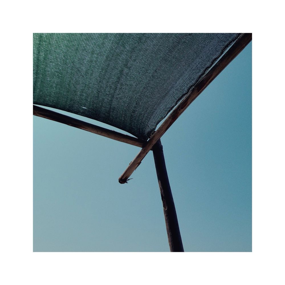 looking at the blue sky through the corner of a rectangular sunshade constructed out of wooden beams and some dark green fabric. Steh image is focusing on the corner of the wooden construction