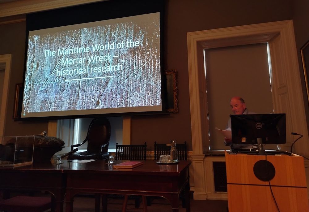 The lecture room in the Society of Antiquaries at Burlington House, London, showing speaker, lectern and the projection screen. The image behind 'The Maritime World of the Mortar Wreck' title slide shows a 13th/14th century graffito of a one-masted ship carved on a pillar in the church of St Mary the Virgin, Hayling Island, Hampshire UK. Photo courtesy of Tom Cousins.