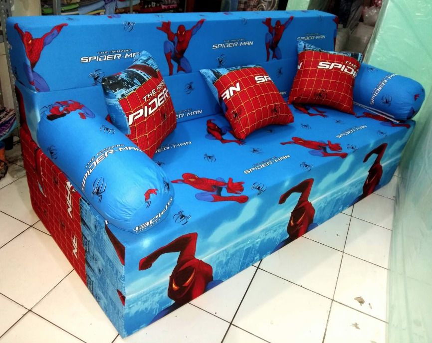 big spider-man couch, looks blocky and spongey, three throw pillows, the red/blue design is sort of upholstered all over the place, repeating images of spider-man doing posts and logos and webbing and new york skyline, couch is on tile in a room