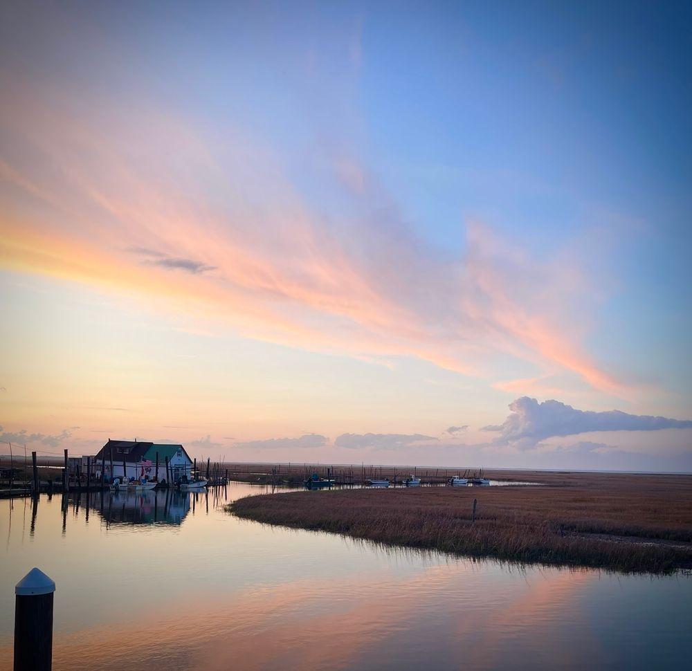 Sunset over a saltwater bay with a solitary house in the distance, surrounded by a mostly empty dock with a couple of boats