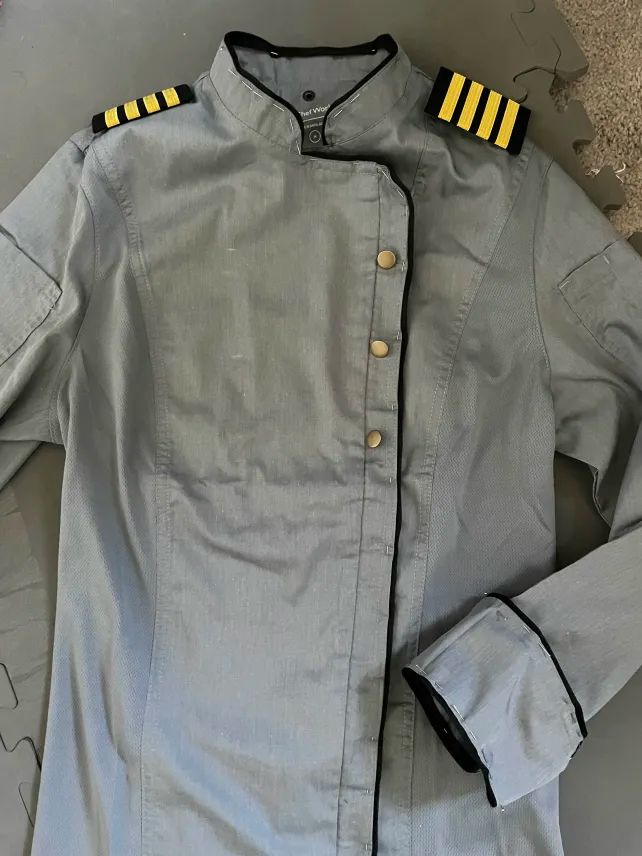 A gray chef's jacket is laid out on the floor. A pair of black-and-gold rectangular epaulets, like those worn by airline pilots, rest on its shoulders. Black piping runs around the collar and down the front closure of the jacket, as well as around the cuffs of the sleeves, visibly pinned in place by many sewing pins. Three brass buttons are laid out on the front closure but not yet sewn in.
