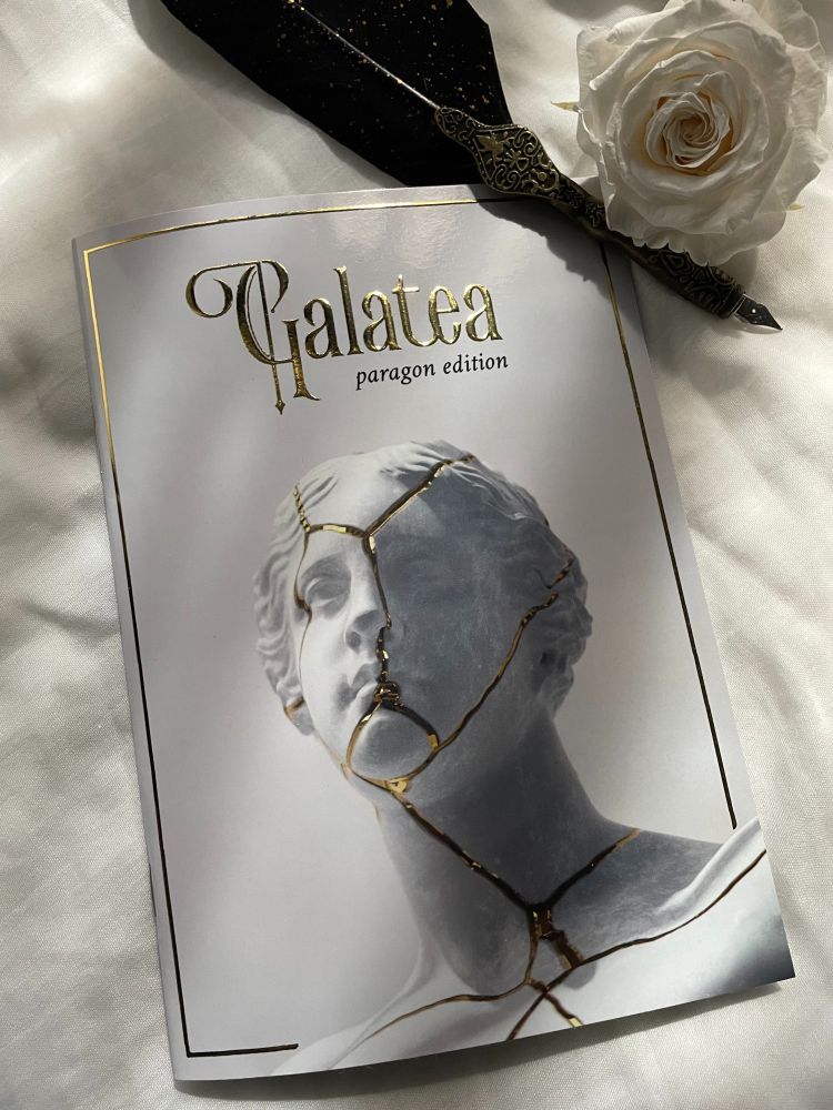 A print zine of Galatea, a solo journaling game by S. Kaiya J., resting on a white linen background next to a quill pen and a white rose.