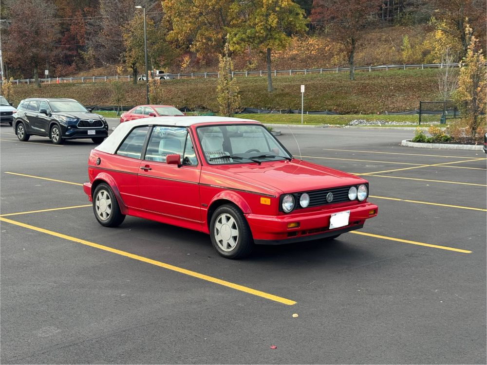A front end shot of a bright red VW Cabriolet convertible with a tapered front end with four circular headlights. It has the slightly boxy, sheet metal look of 80s and early 90s Volkswagens