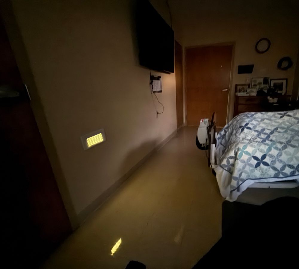 A flash-lit hospital bedroom with a single emergency light the size of an HVAC vent glowing on the floor. The TV and every other light is off in the room while a patient tries to rest under a growing pile of blankets.