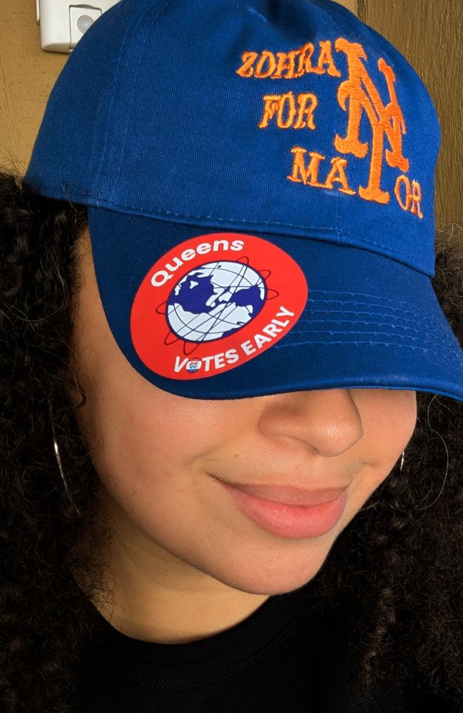 A selfie of a light skinned mixed woman with curly brown hair. She's wearing hoops and a blue baseball cap over her eyes that says "Zohran for NY Mayor" in the style of the NY Mets baseball logo. It has a "Queens Votes Early" sticker on the brim.