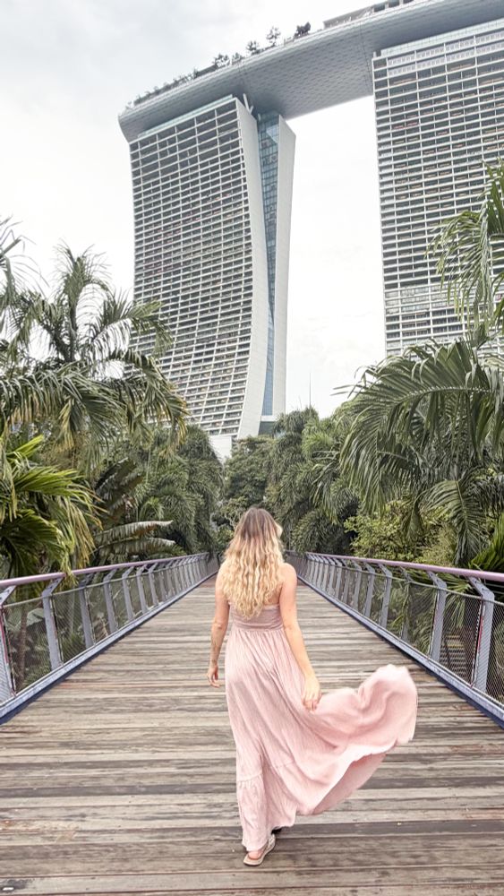 Hübscheste Tochter der Welt mit bestem Herzen der Welt geht auf erhöhten Steg zwischen Palmen auf die weißen Türme der Marina Bay Sands in Singapur zu, links und rechts mit dichten Palmen.