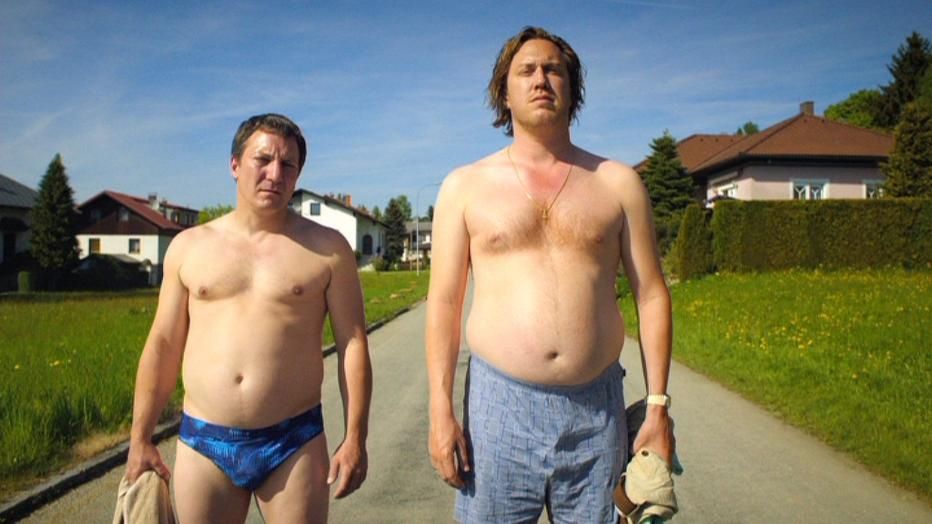 Palfrader und Ofczarek, mit Badehose bekleidet, auf kleiner Straße in Richtung Braunschlag. Links und recgts der Straße eine Wiese und ain paar Häuser mit Hecke. 