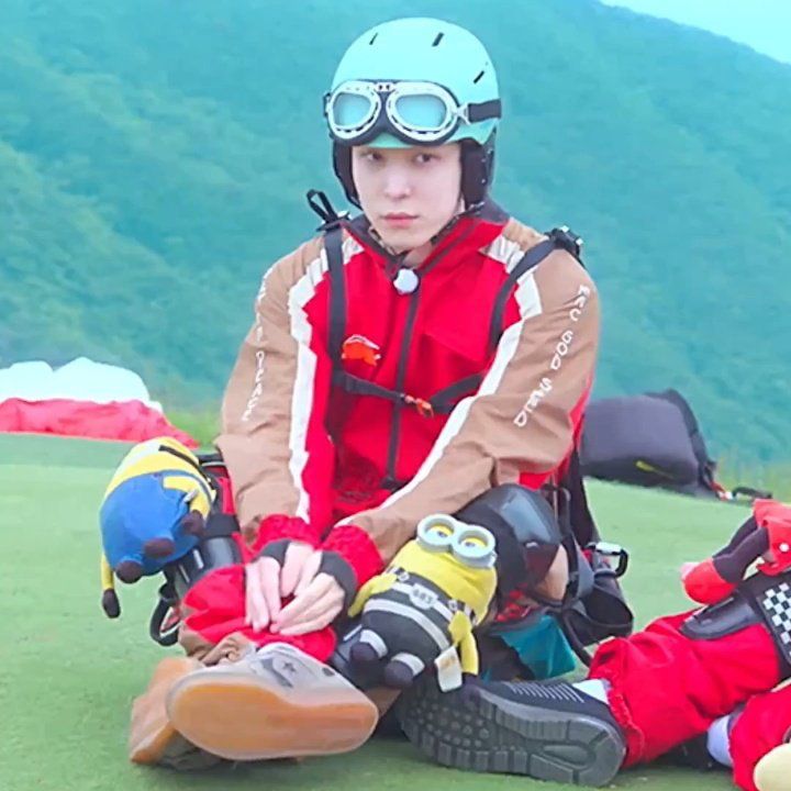 yunho in a paragliding outfit. it’s red and brown. he’s wearing a blue helmet with goggles and has knees pads that are minion plushies. he is pouting. 