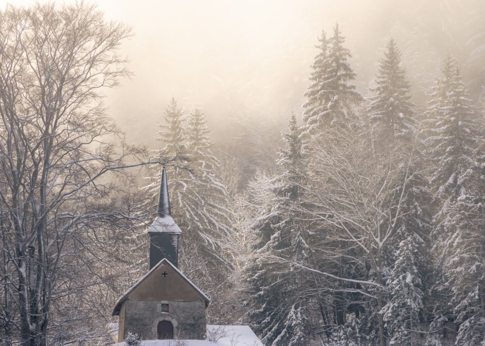 Small chapel in the French alps after a snowfall, with warm yellow morning sunlight diffusing through the clouds