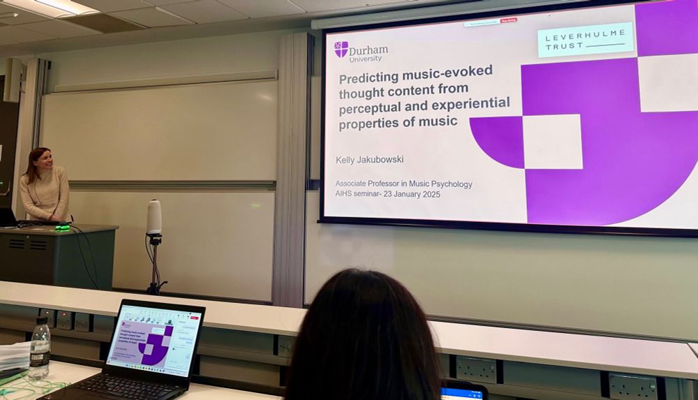 Kelly standing in front of a class talking. A slide shows “Predicting music-evoked thought content from perceptual and experiential properties of music” 