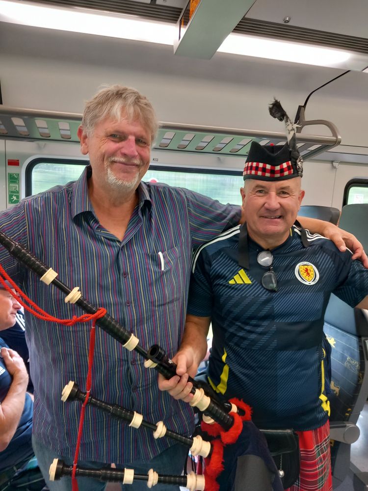Ein älterer Mann in einem gestreiften Hemd legt seinen Arm um einen weiteren älteren Mann im Trikot der schottischen Nationalmannschaft. Er hat eine traditionelle schottische Mütze auf und einen Dudelsack in der Hand. Das Bild ist in einem Bahnwaggon aufgenommen.