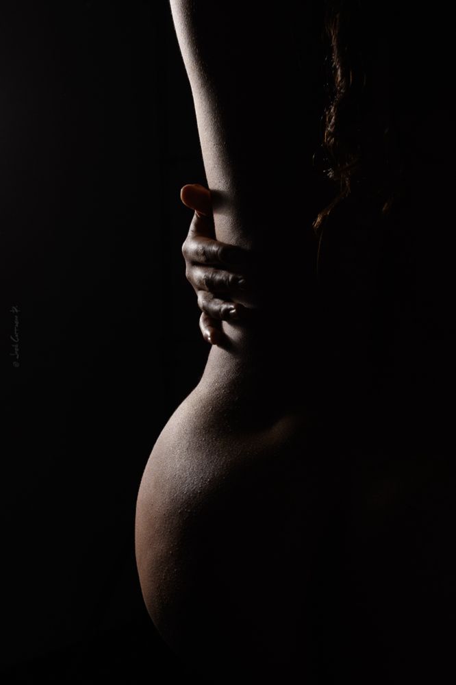 This is a striking and artistic boudoir photograph featuring a close-up of a nude body in dramatic low-key lighting. The composition highlights the natural curves of the subject, with the soft glow of light emphasizing the texture of the skin. A hand gently grasps the waist, adding an element of intimacy and self-embrace. The dark background creates a strong contrast, focusing all attention on the illuminated portions of the body. The image exudes sensuality, elegance, and mystery, making use of shadows and highlights to create depth and emotion.