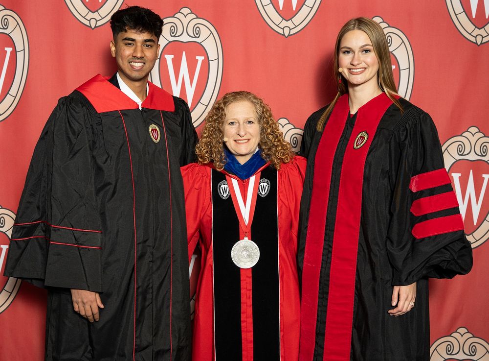 Student speaker Jeeva Premkumar, Chancellor Jennifer Mnookin, and keynote speaker Grace (Stanke) Vanderhei smiling together in full regalia in front of a red background with the University of Wisconsin crest repeated. 