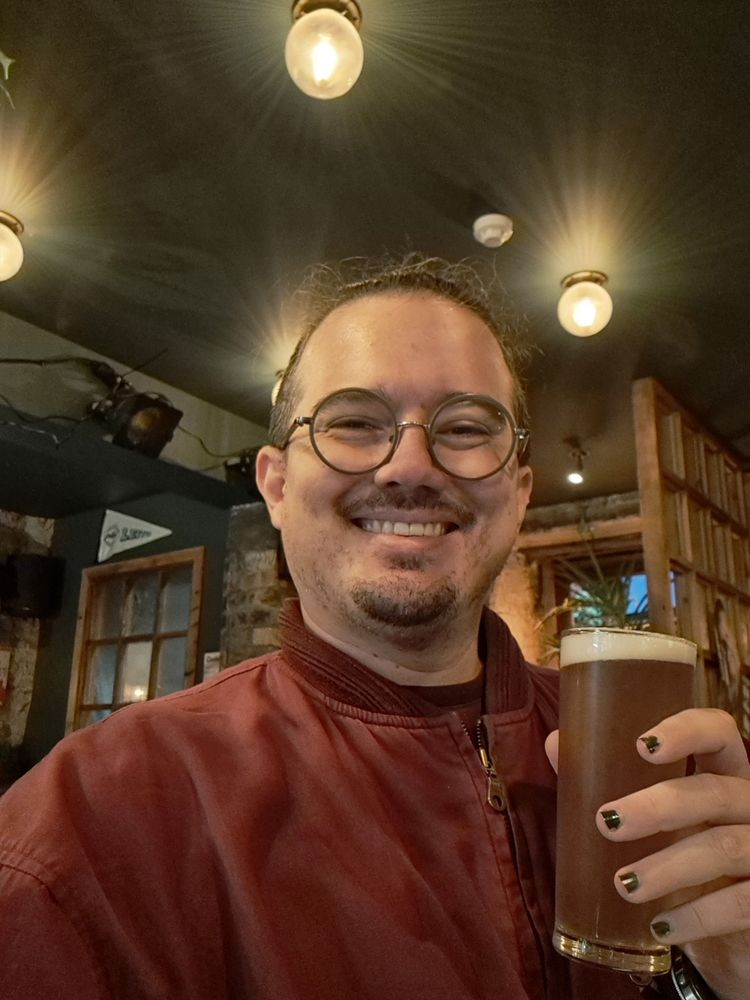 A round faced white guy with goateeand stubble smiles at the camera and raises a half pint glass, wearing a cool red jacket in a warmly lit pub