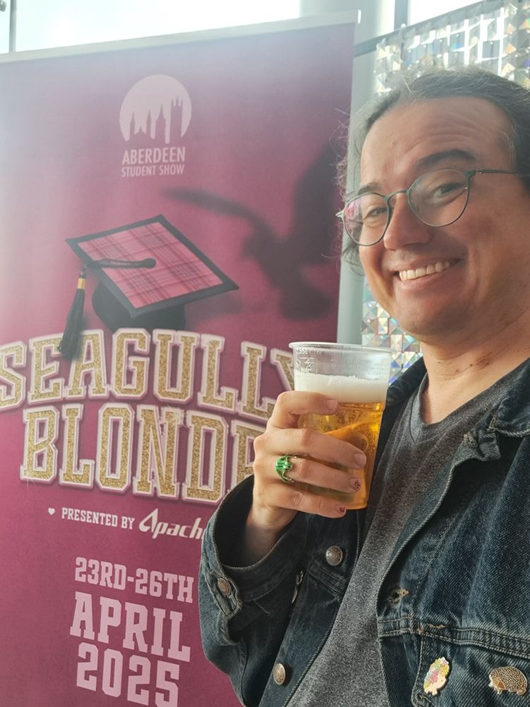 Dave poses in front of a pink poster for the show "Seagully Blonde" wearing their denim jacket and holding a beer