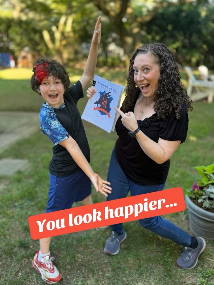 Words on screen: "You look happier..."
Background: a backyard with trees that is mostly blurred out. 
On left in foreground: 10yo smiling boy with wavy brown hair and part of the front of his hair is dyed red. He is wearing a blue swim shirt and shorts with red sneakers and is extending his arms out in joy. 
On right: 42yo mom with long curly brown hair, wearing a black shirt and skinny jeans with white sneakers. She is holding and pointing to a manuscript that has a shield with the letters "MCC" and the title "The Mythical Creature Conservatory." They are joyous. 