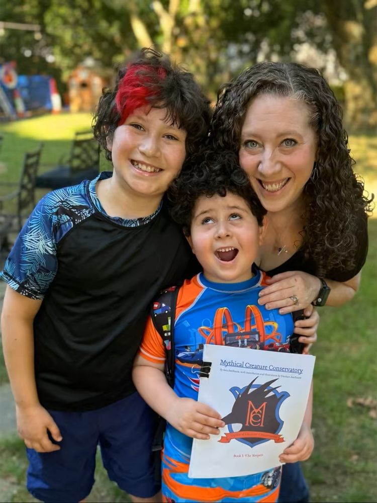 Background: a backyard with trees that is mostly blurred out. 
Foreground is a mom with her two children. 
On left: 10yo smiling boy with wavy brown hair and part of the front of his hair is dyed red. He is wearing a blue swim shirt and shorts
On right: 42yo mom with long curly brown hair, wearing a black shirt with her arms around the two children. 
In front of them is a 6yo boy with curly dark hair wearing an orange and blue swimsuit and shirt. He is looking up and making a silly happy face. 
The 6yo is holding a manuscript that has a shield with the letters "MCC" and the title "The Mythical Creature Conservatory."