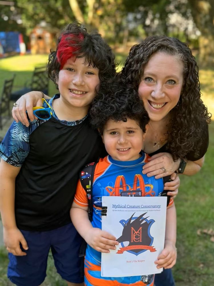 Background: a backyard with trees that is mostly blurred out. 
Foreground is a mom with her two children. 
On left: 10yo smiling boy with wavy brown hair and part of the front of his hair is dyed red. He is wearing a blue swim shirt and shorts
On right: 42yo mom with long curly brown hair, wearing a black shirt with her arms around the two children. 
In front of them is a 6yo boy with curly dark hair wearing an orange and blue swimsuit and shirt. 
The 6yo is holding a manuscript that has a shield with the letters "MCC" and the title "The Mythical Creature Conservatory." They are smiling