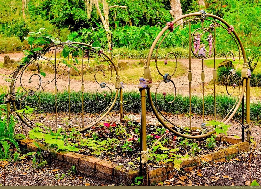 This brick flower bed has a full-size, traditional, ornate, metal bed frame, with floral flourishes and cherubs. There is lots of greenery, with some ivy growing up around the bed posts, and some red flowers in the bed (looking very comfortable).   :) 