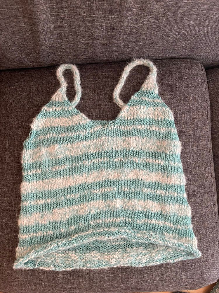 A blue and white horizontally-striped knit tank top is displayed on a grey couch cushion. The white stripes alternate thick and thin. The tank top is a v-neck with icord straps.  