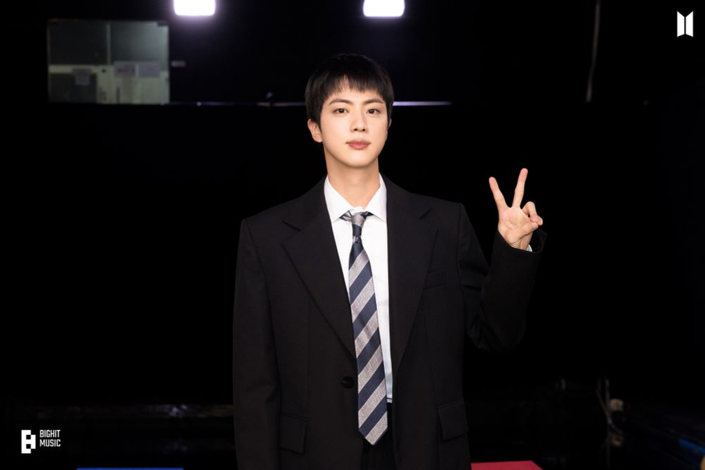 Jin of BTS wearing a formal black suit, a striped tie, and a white shirt stands against a dark background with bright lights above him. He holds up a peace sign with his fingers and has a calm expression. The logo in the bottom-left corner reads "BigHit Music."
