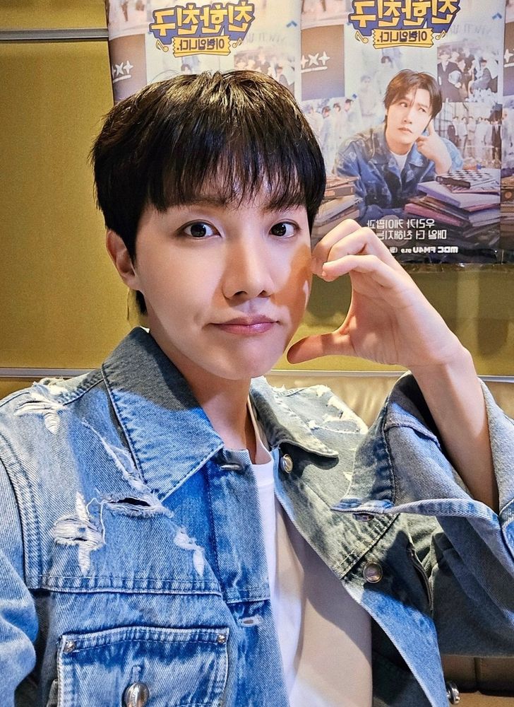 J-hope of BTS wearing a distressed denim jacket and a white shirt is taking a selfie. He's forming a heart shape with his hand near his face. In the background, there are colorful posters featuring another Korean man in a similar denim outfit, with text in Korean.

