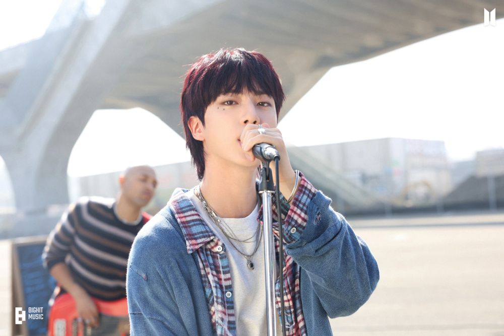 Jin of BTS singing into a microphone, wearing a plaid shirt and denim jacket, with a bridge structure and a bass player blurred in the background.



