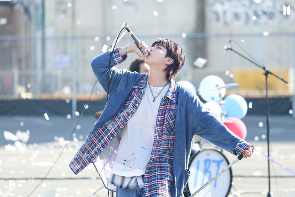 Jin of BTS passionately singing into a microphone, wearing a layered outfit of a plaid shirt and a denim jacket, with balloons and confetti in the background.


