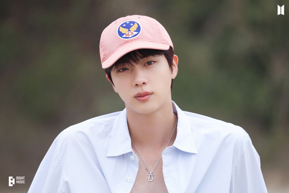 Jin of BTS wearing a pink cap with a logo, a light blue unbuttoned shirt, and a silver necklace, standing outdoors with a blurred green background.