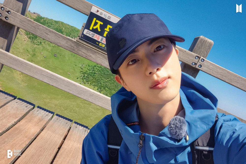 Jin of BTS dressed in a blue hiking jacket and a navy cap takes a selfie outdoors on a wooden structure. The background features a vibrant green grassy landscape under a clear blue sky. A sign with text is visible behind him, adding detail to the scene. The logo in the bottom-left corner reads "BigHit Music."