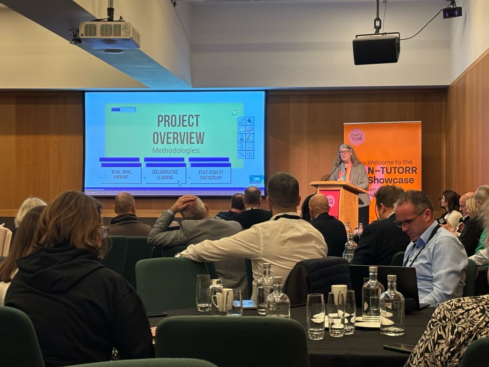 Woman speaking at top of room. Slide visible says ProjectOverview. Audience visible in foreground 