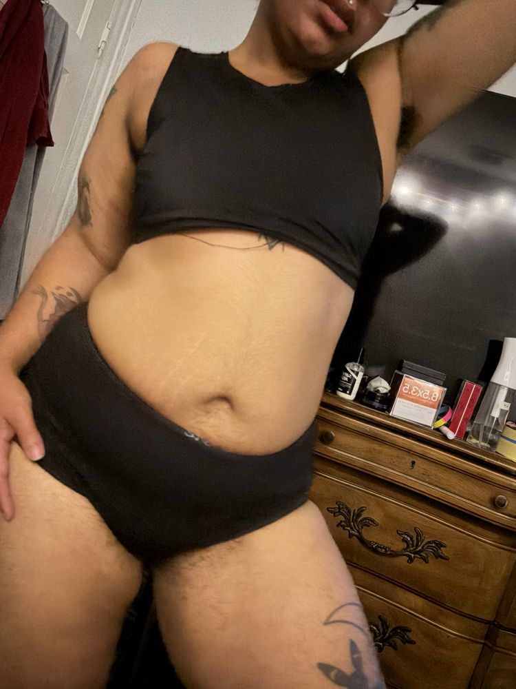 A photo of Cornelius standing in their room with black panties and a black binder, their hand is on their hip and the other is behind their head