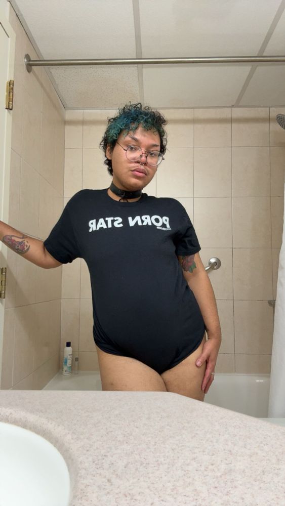 A photo of Cornelius standing in a bathroom of a hotel room, without any pants. He’s wearing a black shirt that says Porn Star in while bold capital letters. His hand is leaning on the wall and the other is on his thigh