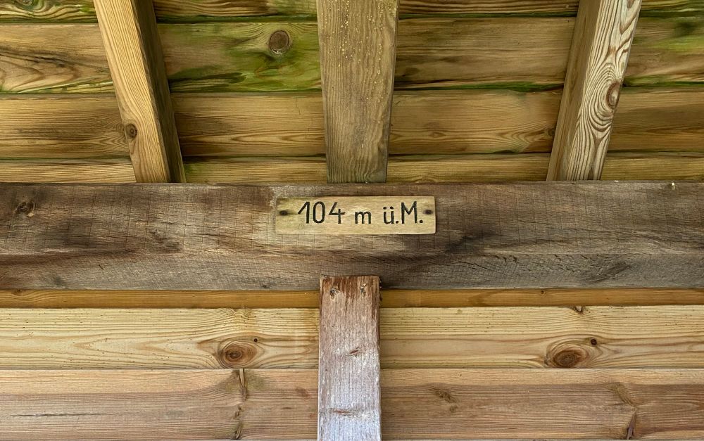 Holzschild mit eingeschnitztem Schriftzug »104 m. ü.M.« an Holzhütte 