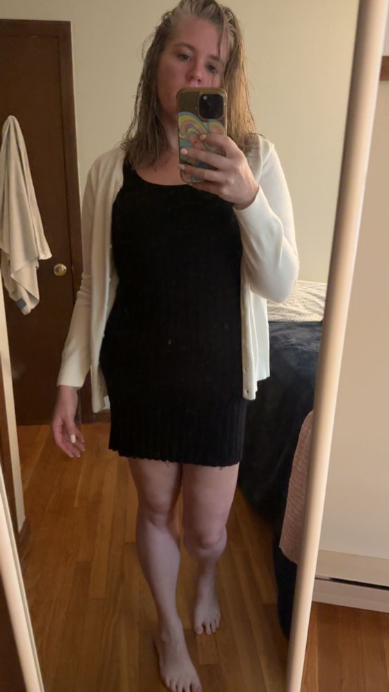 Dahlia with bare feet in front of a mirror wearing a black dress and white sweater 