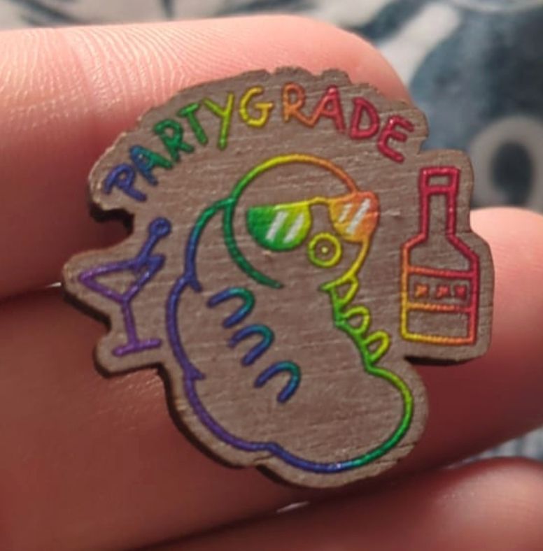 small wooden pin with a rainbow tardigrade with a martini glass and alcohol bottle wearing sunglasses with the text PARTYGRADE above.