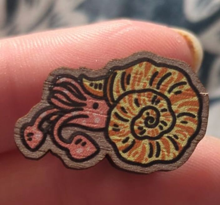 small wooden pin of our mascot extinct invertebrate, a yellow and orange shelled ammonite with pink tentacles.