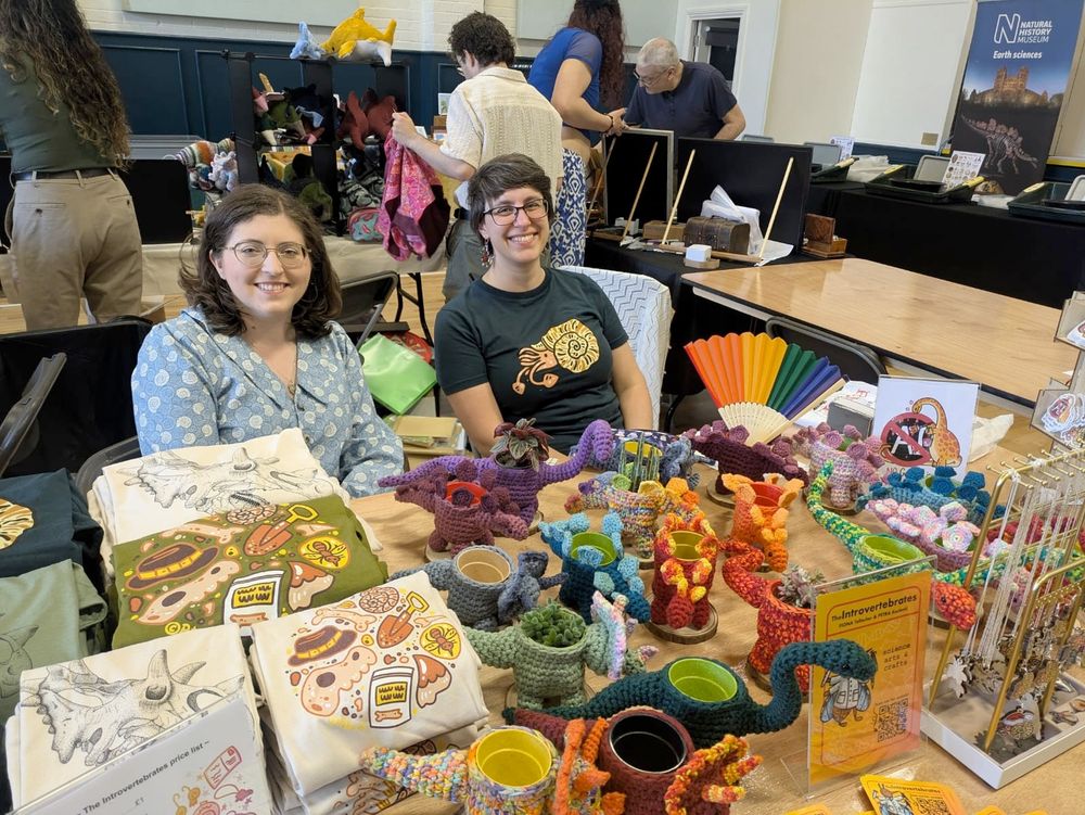 A hall full of people setting up tables, two people are sitting down onr with a light blue ammonite blouse the other dark blue cartoony ammonite, the table has multicolor dinosaur crochet planters, paleonthology tshirts, a rack with jewelry, an anti ai sign.