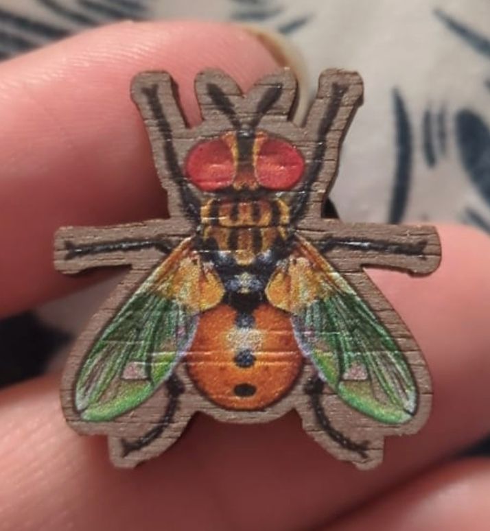 small wooden pin of a ladybug fly, a fly with bright red eyes, green yellow wings, and a dark orange abdomen with three black dots making it look like a ladybug.