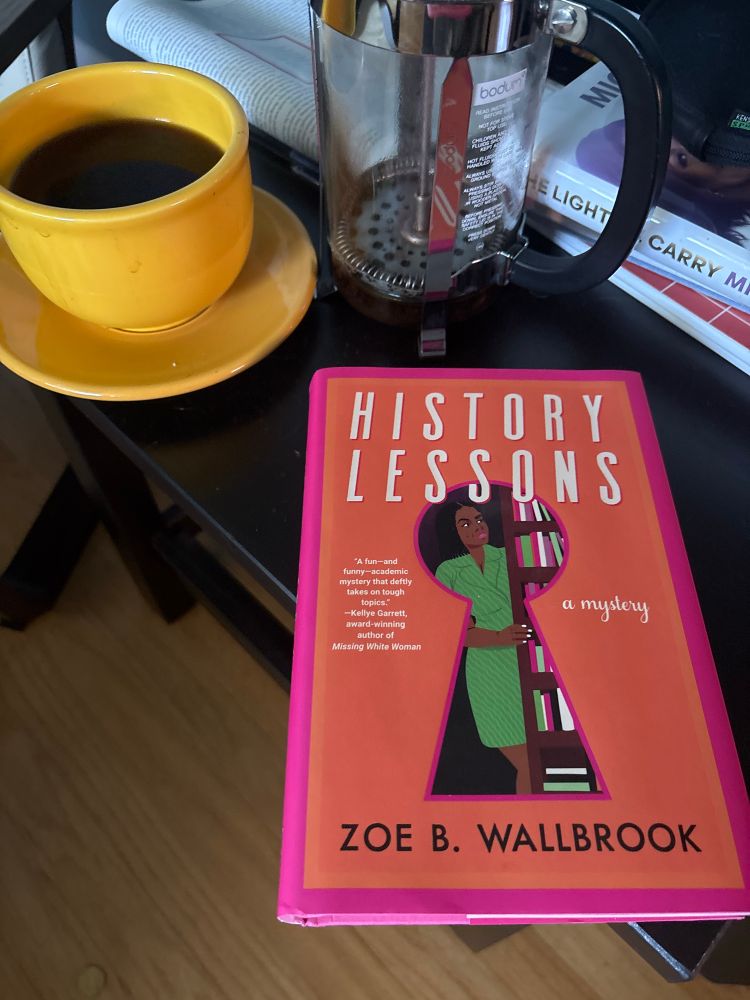 The orange cover of History Lessons by Zoe B Wallbrook, showing a black woman in a green dress outlined in a keyhole as she peeks around a bookcase. The book sits on a table next to a yellow coffee cup. 