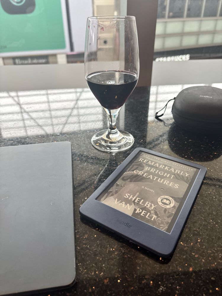A blue Kindle showing the cover of Remarkably Bright Creatures by Shelby Van Pelt sits on a black granite countertop next to a glass of wine and in front of a large window in an airport lounge.